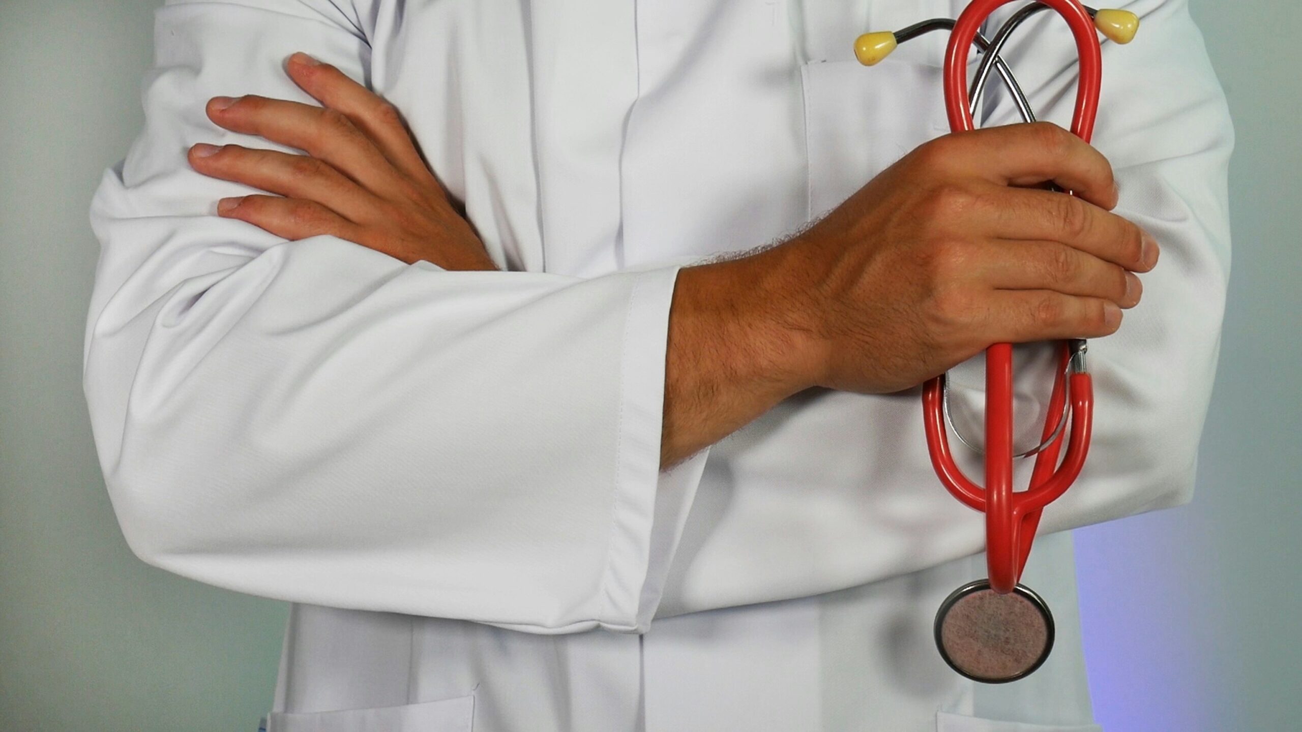 A healthcare professional in a crisp white medical coat stands with arms crossed, holding a red stethoscope in one hand against a clean, light-colored clinical background. The image conveys professionalism and medical expertise, making it ideal for provider spotlight posts, general healthcare content, or promoting the practice's medical services and professional care standards.