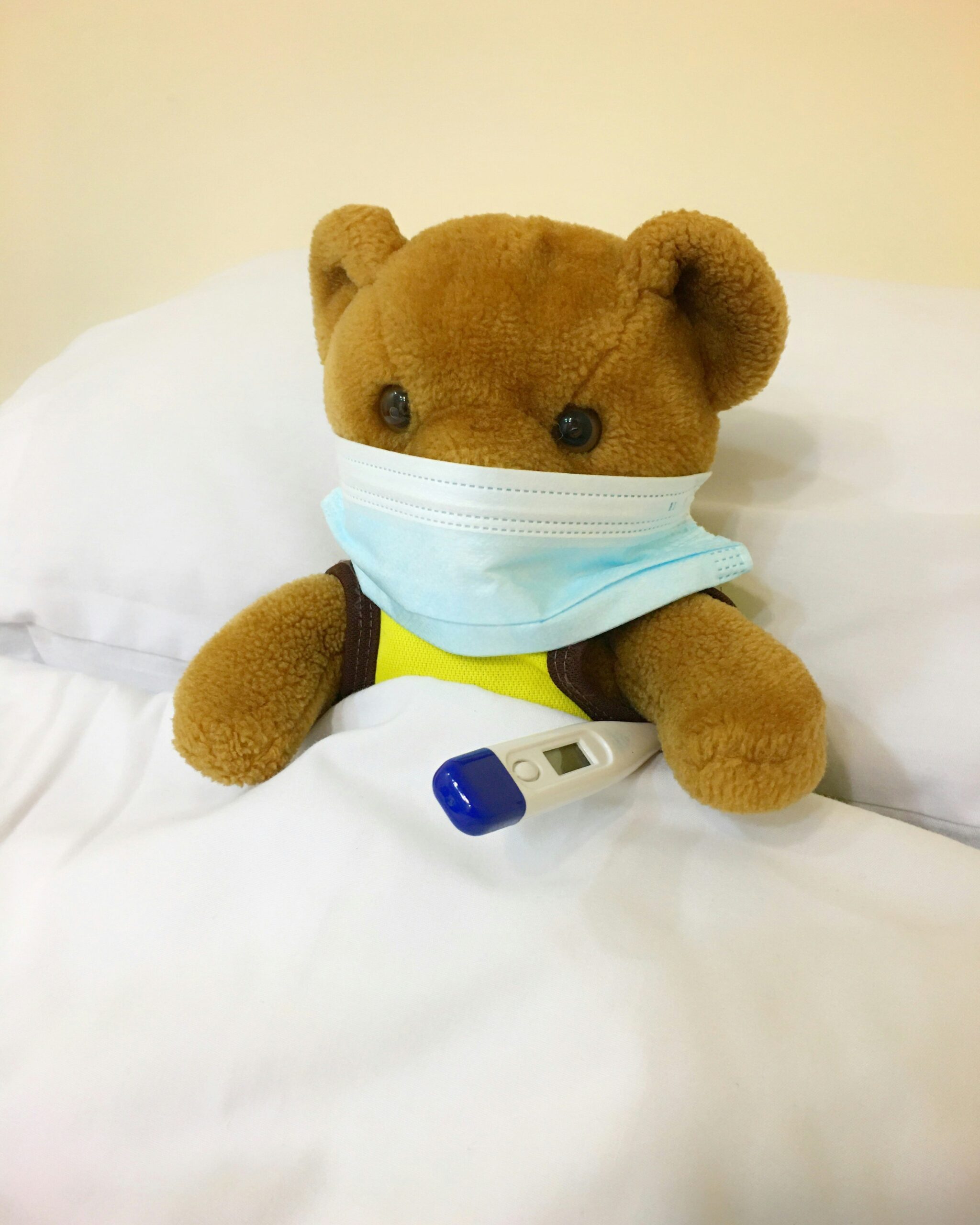 A brown teddy bear wearing a yellow shirt and a light blue surgical face mask pulled down around its chin sits propped up against white bedding, with a digital thermometer tucked in front of it. The playful, cozy setup mimics a sick child resting in bed and conveys a gentle, reassuring tone. This image would work well for social posts about pediatric care, same-day appointments for acute illness, or seasonal flu and cold-season health tips.